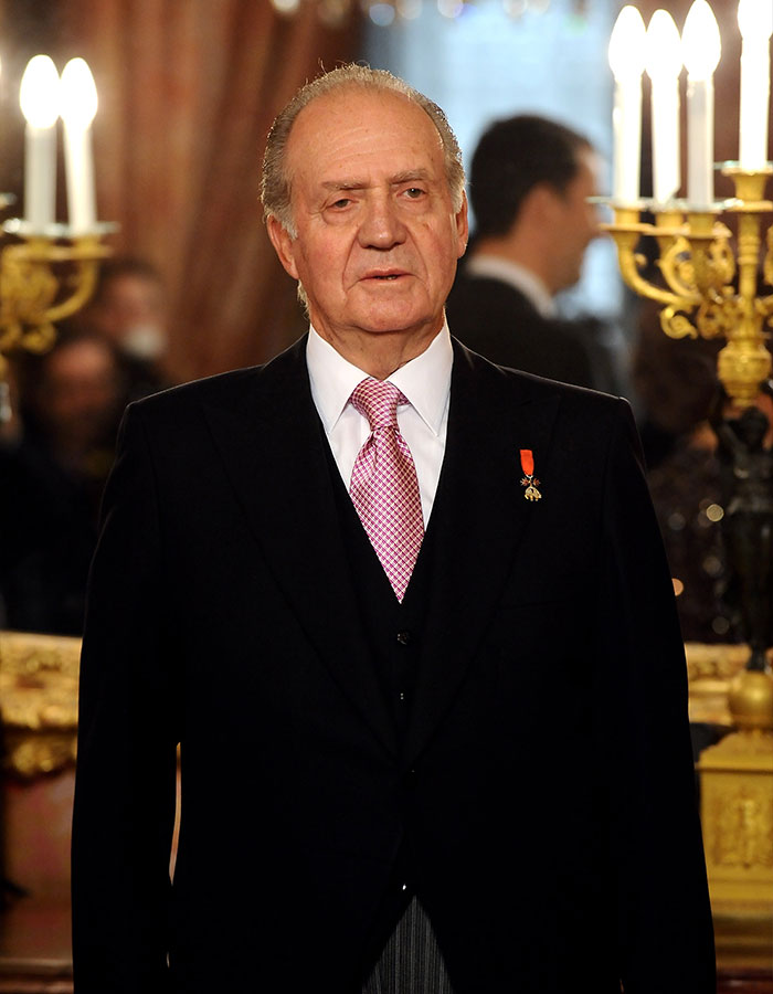 Former King Juan Carlos dressed formally at an indoor event with candles and people in the background.