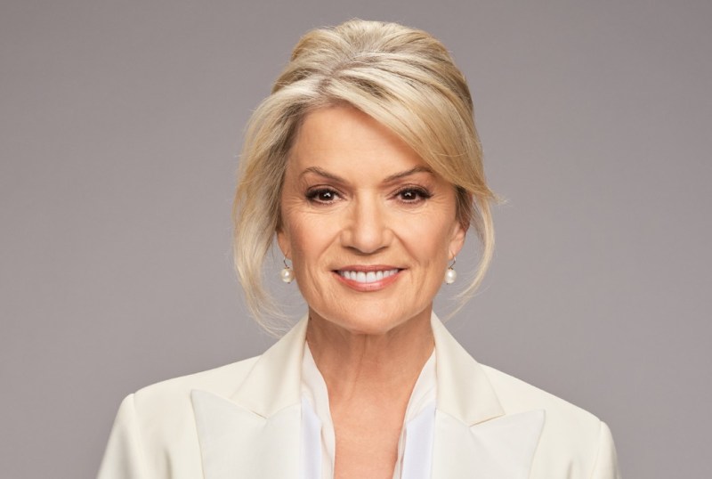 Portrait of a smiling woman with blonde hair styled in soft waves, wearing a white blazer and pearl earrings, set against a neutral background.