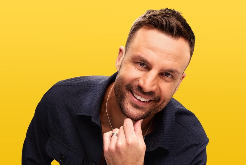Smiling man in a navy shirt with a textured pattern, posing against a bright yellow background, showcasing a friendly and approachable demeanor.