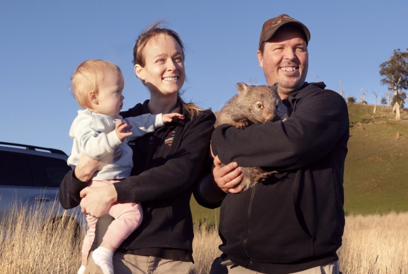 A smiling couple holding a baby and a wombat in a rural Australian setting, with a grassy hill and blue sky in the background. The woman wears a light blue sweater, while the man is dressed in a black jacket and cap. The family enjoys a moment outdoors, showcasing Australia's unique wildlife.