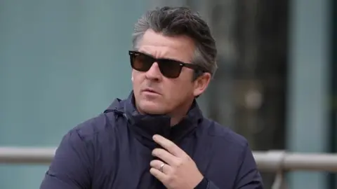 PA Joey Barton has dark greying hair and wears sunglasses. He is wearing a navy blue jacket