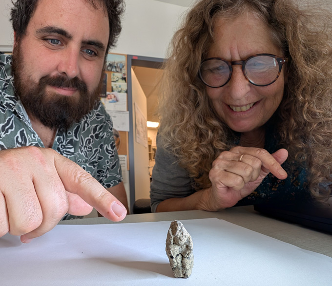 A man points to a tiny clay figurine that sits on top of a sheet of white paper as a woman looks on. 