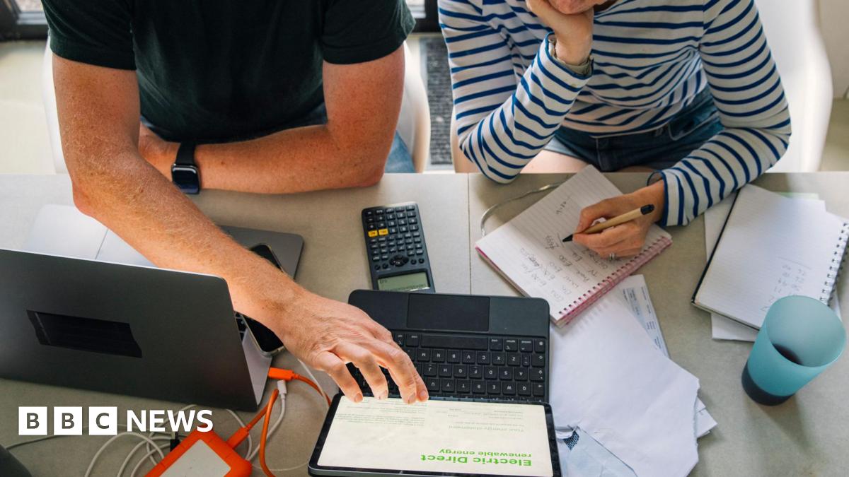 A couple sit at a large table in front of a laptop with a calculator and notebooks. They look to be going over a budgets and finances. The man is dressed in a dark t-shirt while the woman has a long-sleeved striped top.