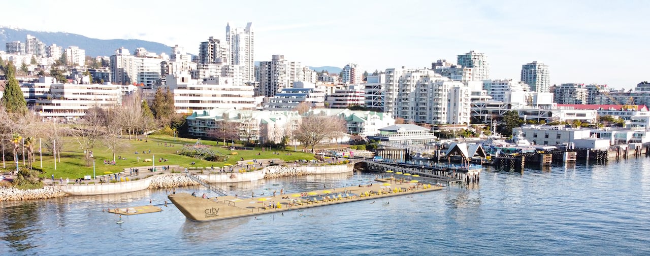 A wooden, floating structure in the ocean in front of a park with city buildings behind it.