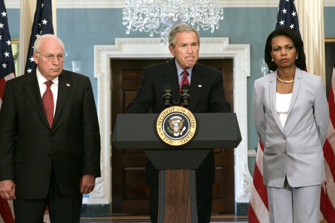 Two cleanshaven Caucasian men in suit and tie are shown standing indoors, next to a dark-complected woman in a pantsuit.