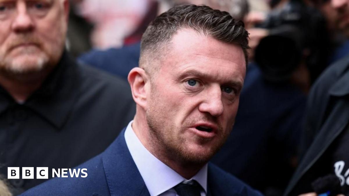 Image shows Stephen Yaxley-Lennon, known as Tommy Robinson, wearing a navvy blue suit, shirt and tie stands outdoors speaking while surrounded by a crowd of people which is slightly out of focus
