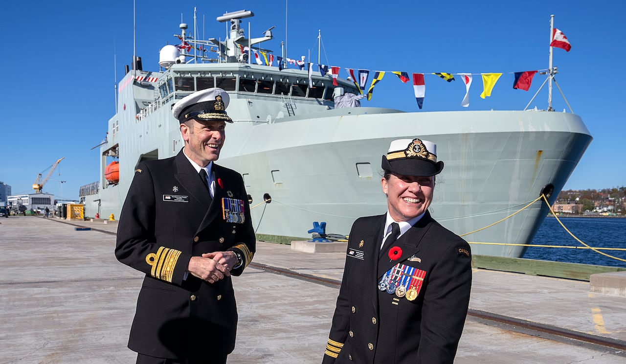 A man and a woman, both in military uniforms, smile while standing outside near a large ship.