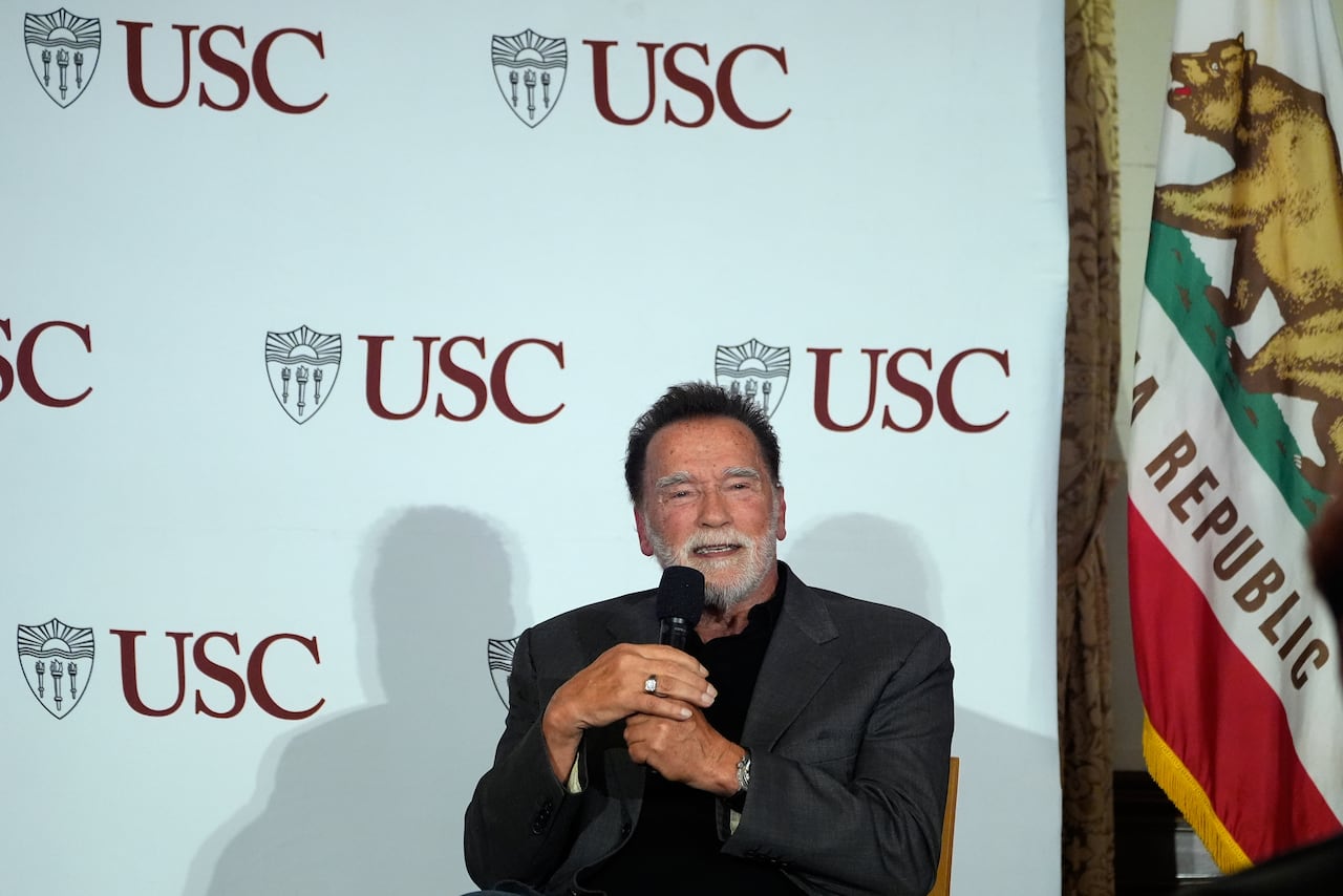 A man with dark hair and a white beard wearning a suit jacket and collared shirt speaks while seated and holding a microphone.