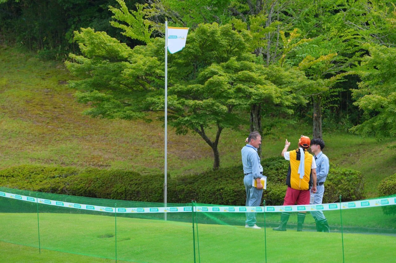 Three people on a golf course point toward a forested area surrounding the course.
