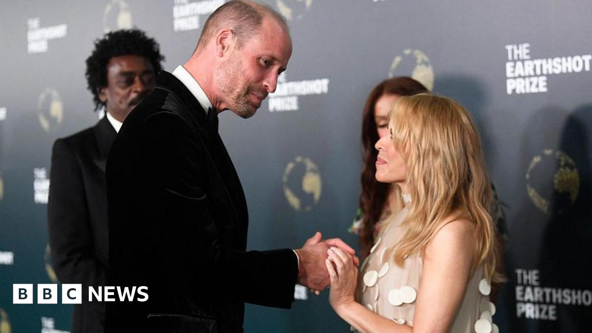Prince William smiles and wears a black velvet jacket as he touches hands with a Kylie Minogue, who is wearing a beige dress with white disc adornments
