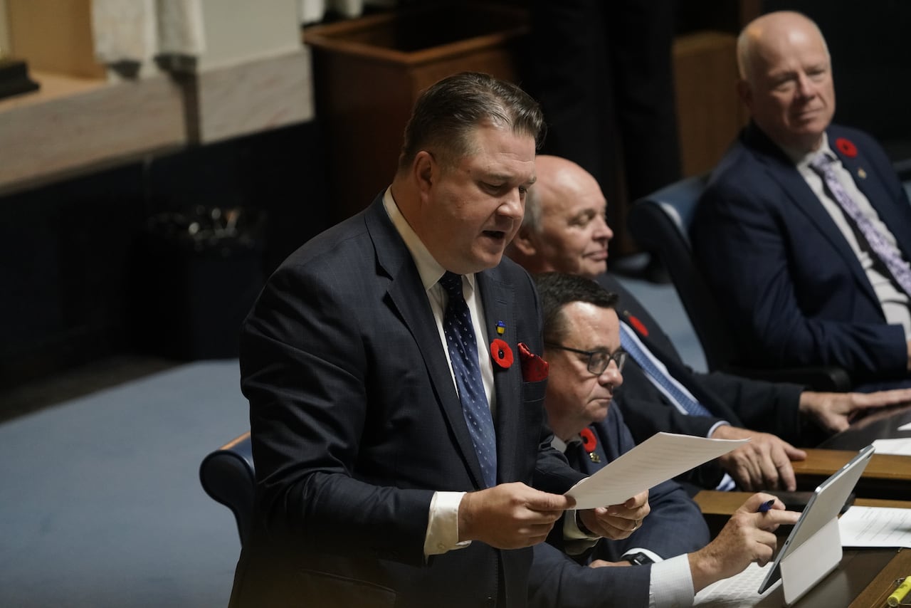 A man in a suit with a poppy on it holds a paper while speaking during a government legislative session.