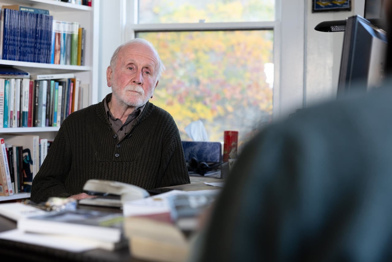 An older man sits at his desk in front of a bookshelf with a neutral expression on his face. Fall coloured leaves are seen out the window behind him.