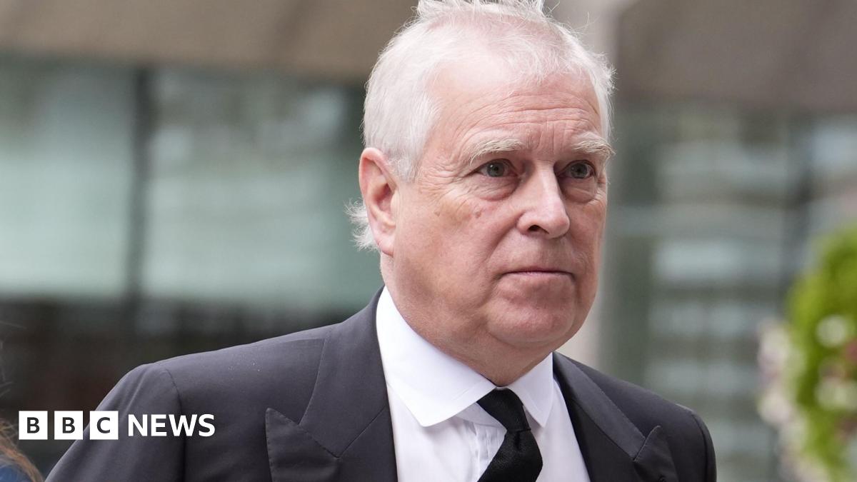 Andrew outside wearing a dark charcoal suit and a black tie, his white hair blowing in the wind - he has a neutral expression on his face