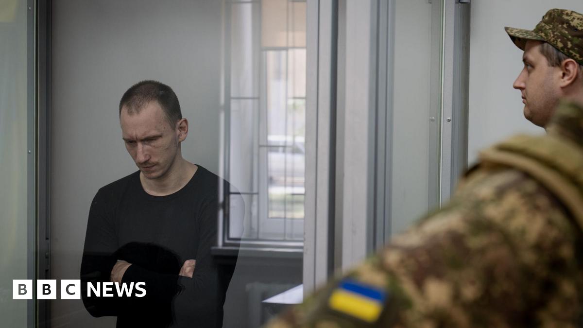 Dmitry Kurashov, who has short dark hair and is wearing a black jumper, stands in the dock with his arms folded, looking down, surrounded by Ukrainian guards.