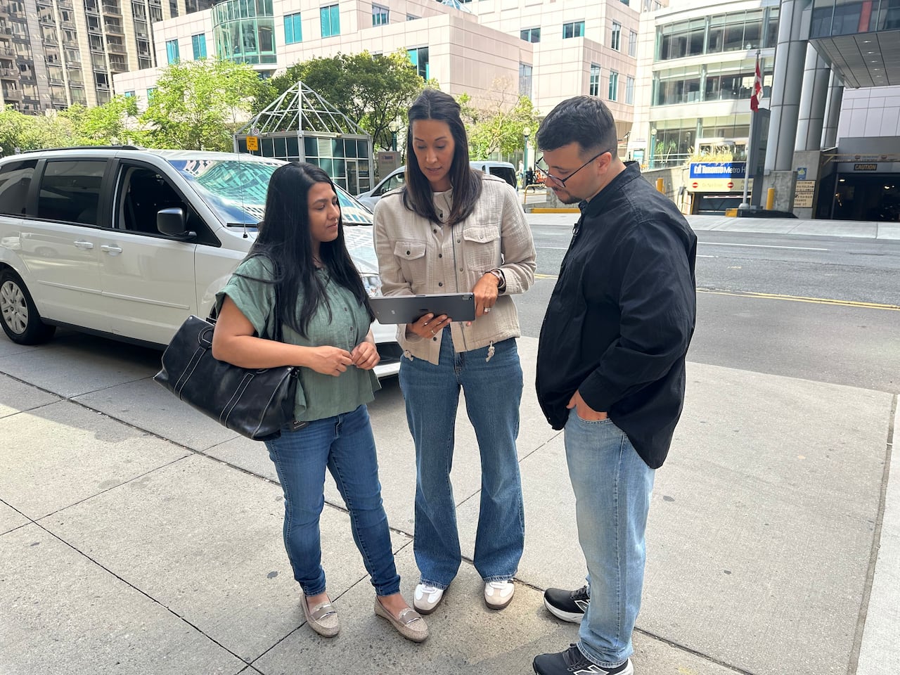 Three people stand and look at a tablet on the sidewalk with a van parked behind them.