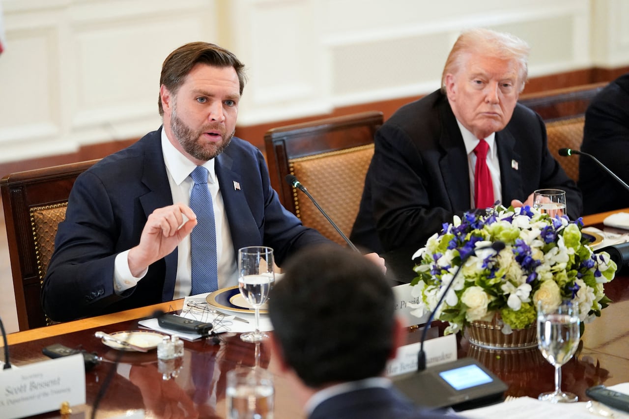 A bearded brown haired man in a suit and tie speaks while seated at a table. Next to him is an older cleanshaven man.