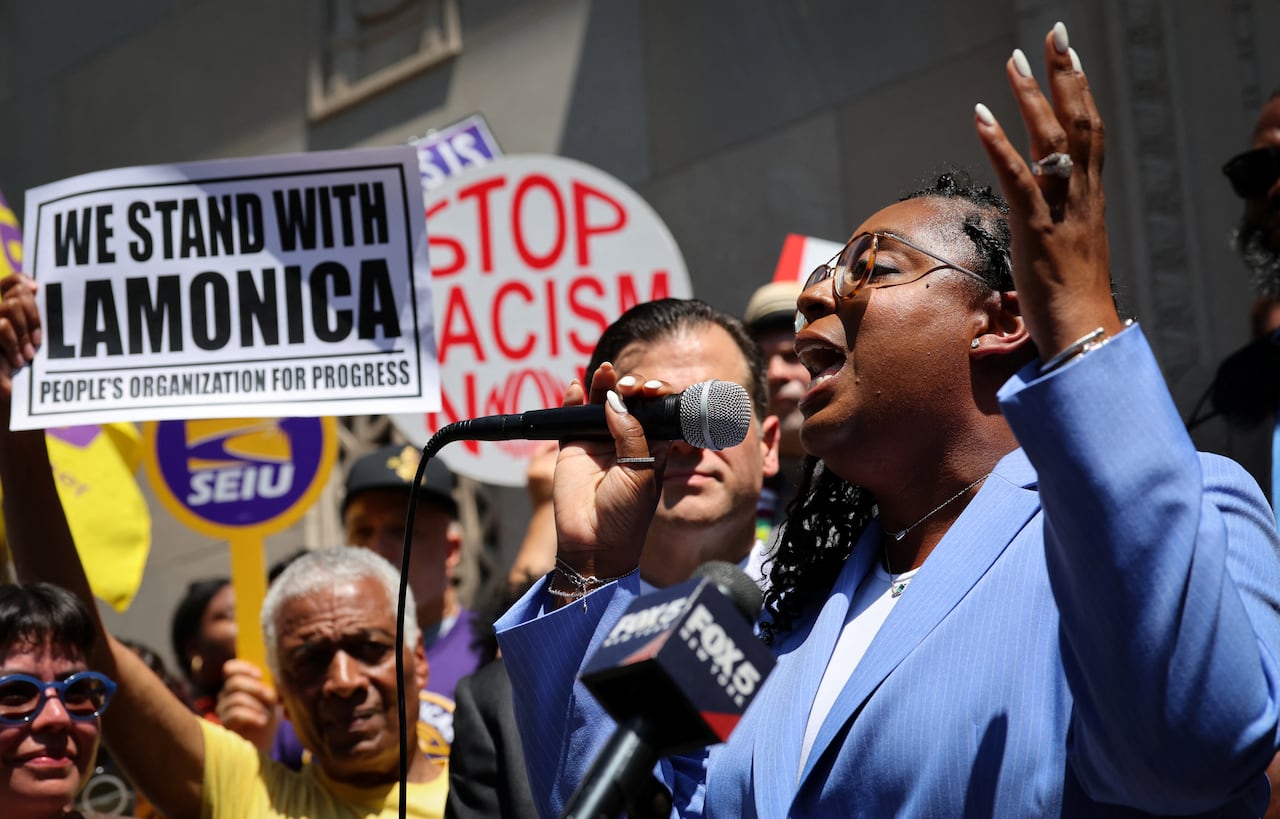A dark-complected, bespectacled woman wearing a light blue blazer is shown in profile speaking into a microphone at an outdoor demonstration.