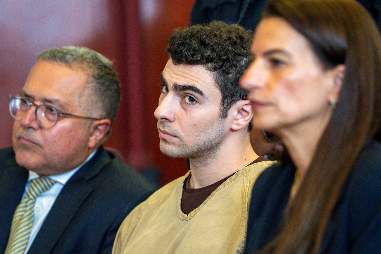 A dark haired man with some stubble on his face turns his head as he sits while wearing a jumpsuit between a man and a woman.