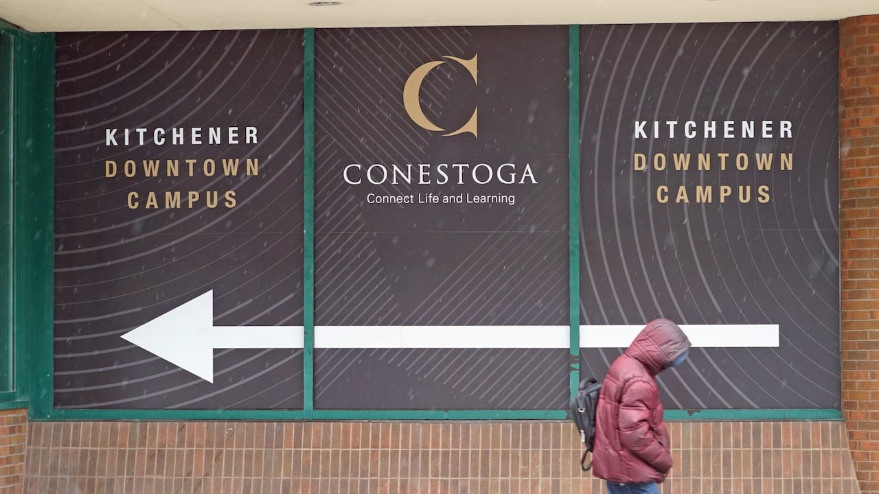 A man wearing a coat walks past a Conestoga College sign.