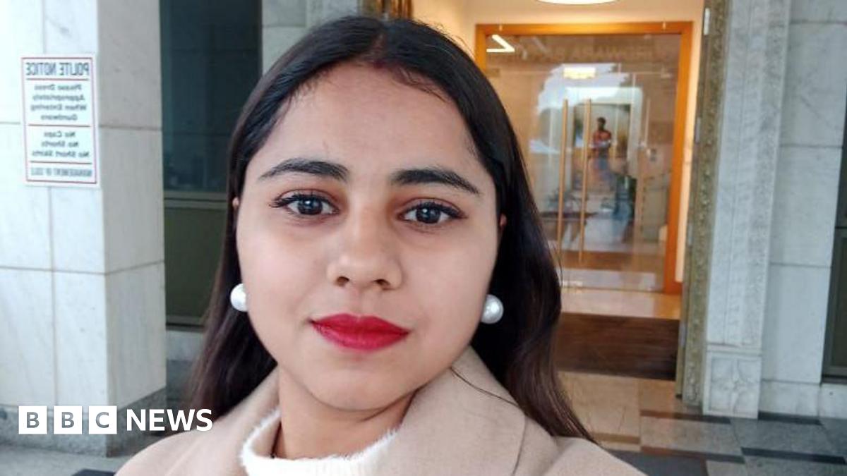 Harshita Brella looks into the camera for a selfie while standing outside a building. Her hair is open, and she is wearing bright red lipstick and pearl drop earrings.