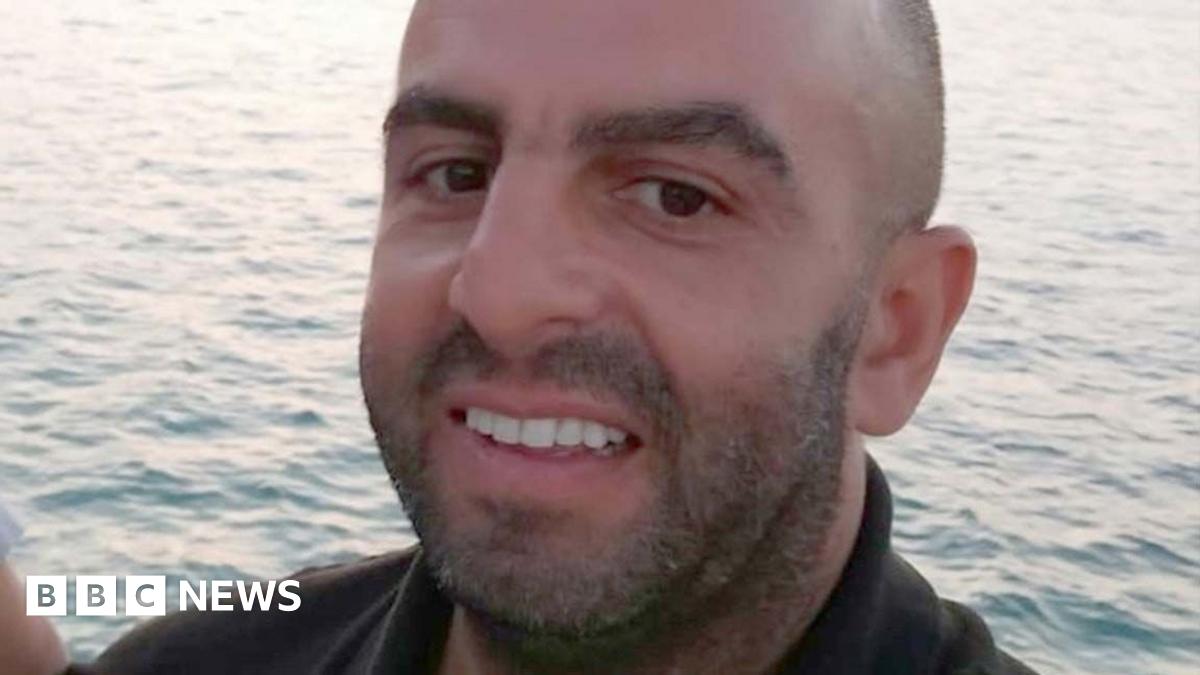 Samir Zitouni is smiling at the camera in a close-up shot of his face. He has a moustache and a beard.