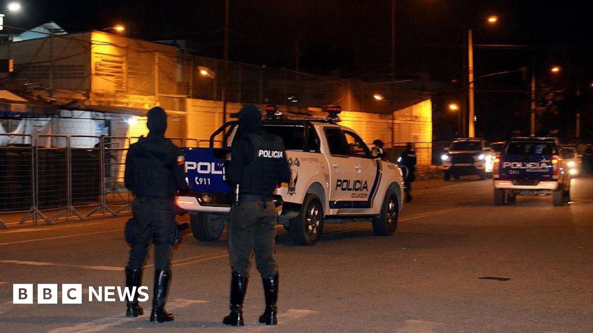 Two police officers stand with their backs to the photographer in front of El Oro prison in the city of Machala, in Ecuador. Behind them, a police pick-up truck and several other police cars can be seen.