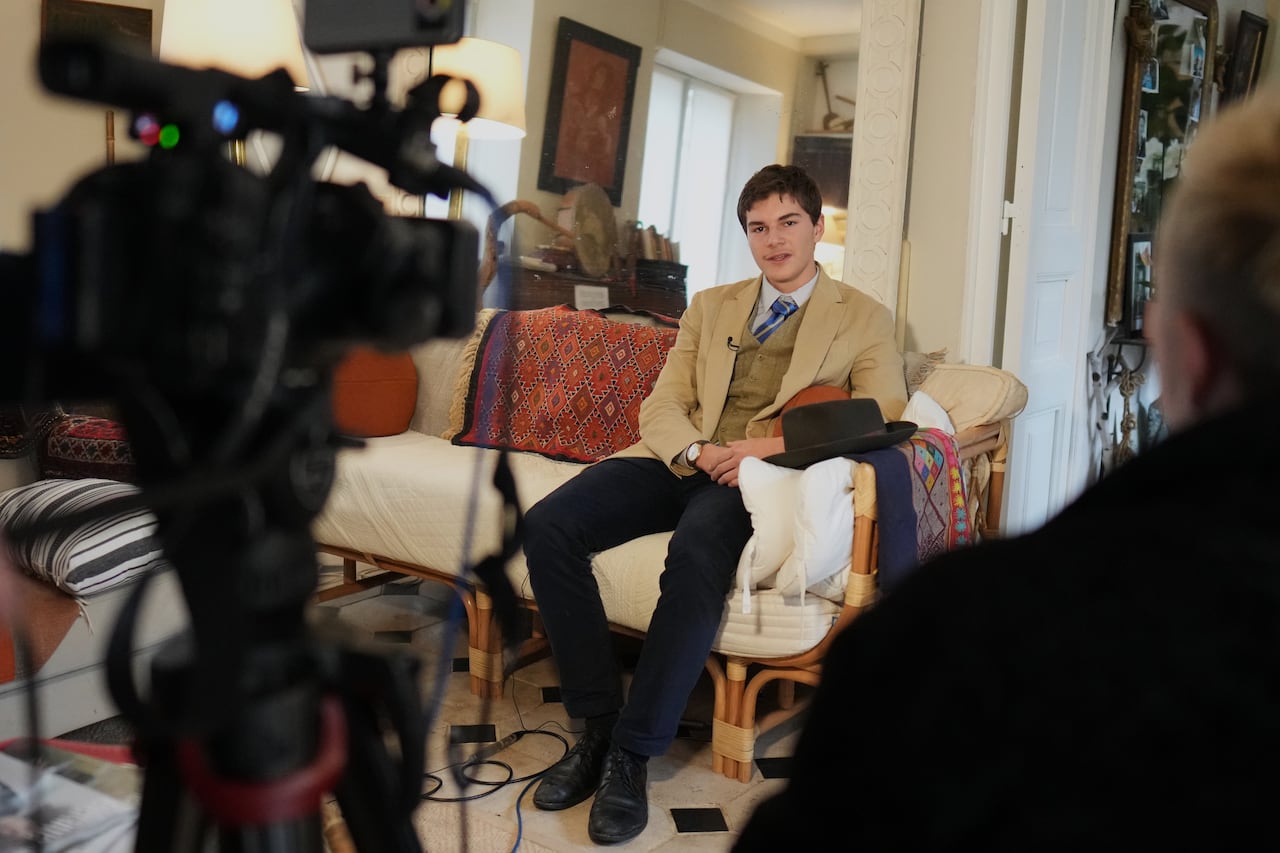 A man in a suit and fedora sits on a  couch and speak to cameras