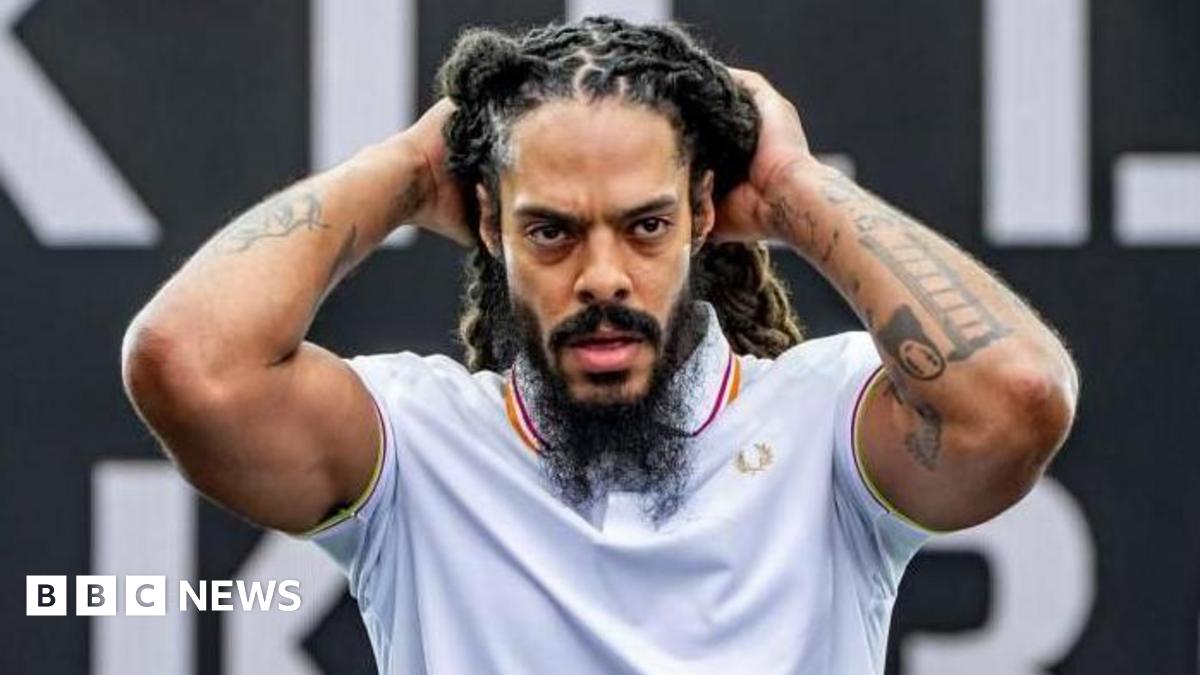 A man performing on stage during a Bob Vylan set.  He is wearing a white polo shirt and his hands are touching his head. He has a dark beard and dreadlocks and tattoos on both his arms.