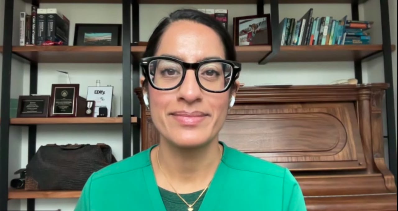 A woman with black hair, pulled back, and black glasses is wearing green scrubs. A piano and shelves containing photos and books are in the background