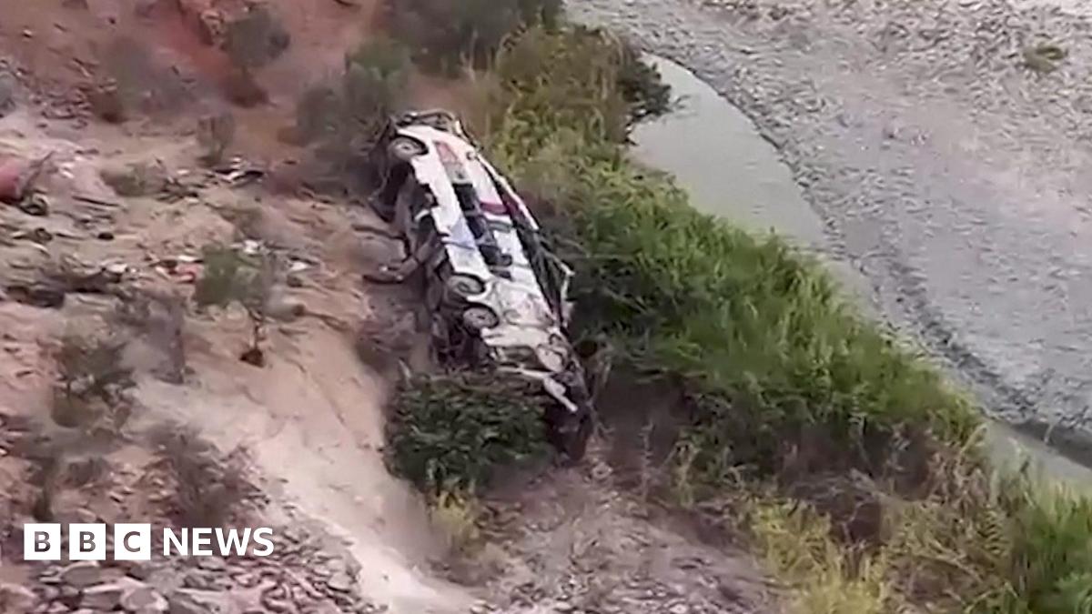 A frame from a video showing a wrecked passenger bus at the bottom of a deep ravine. The terrain is rocky and sandy with a shallow river snaking across the bottom of the ravine. The bus looks utterly destroyed.