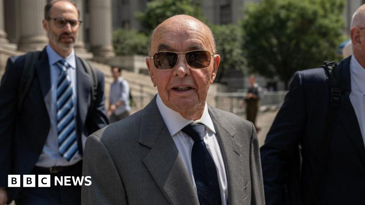 Joe Lewis stands outside a courthouse and wears a grey suit with a black tie and sunglasses.
