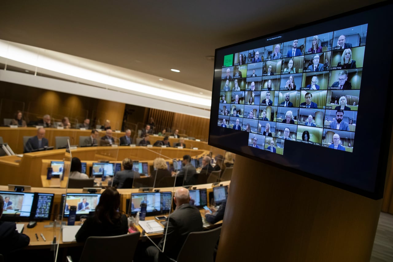 Metro Vancouver elected officials meet around a table on the left while a TV screen showing virtual attendees is on the right