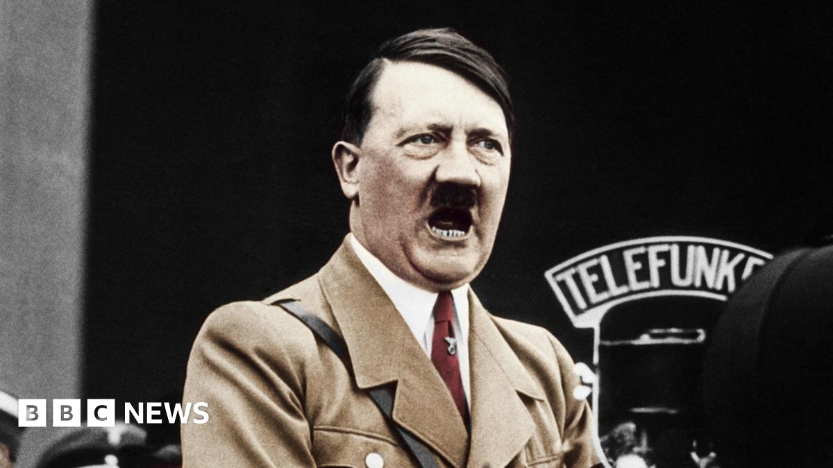 A colour treated photo of Hitler speaking into a big microphone. He's wearing a light brown jacket and red tie.
