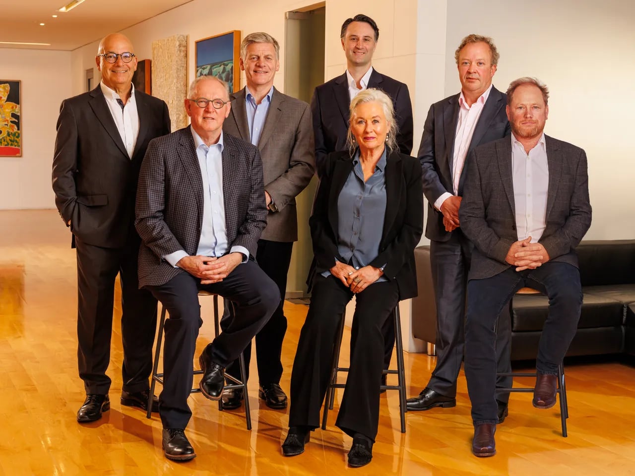 A group of people in suits standing and sitting in two rows, in a room with art in the background.