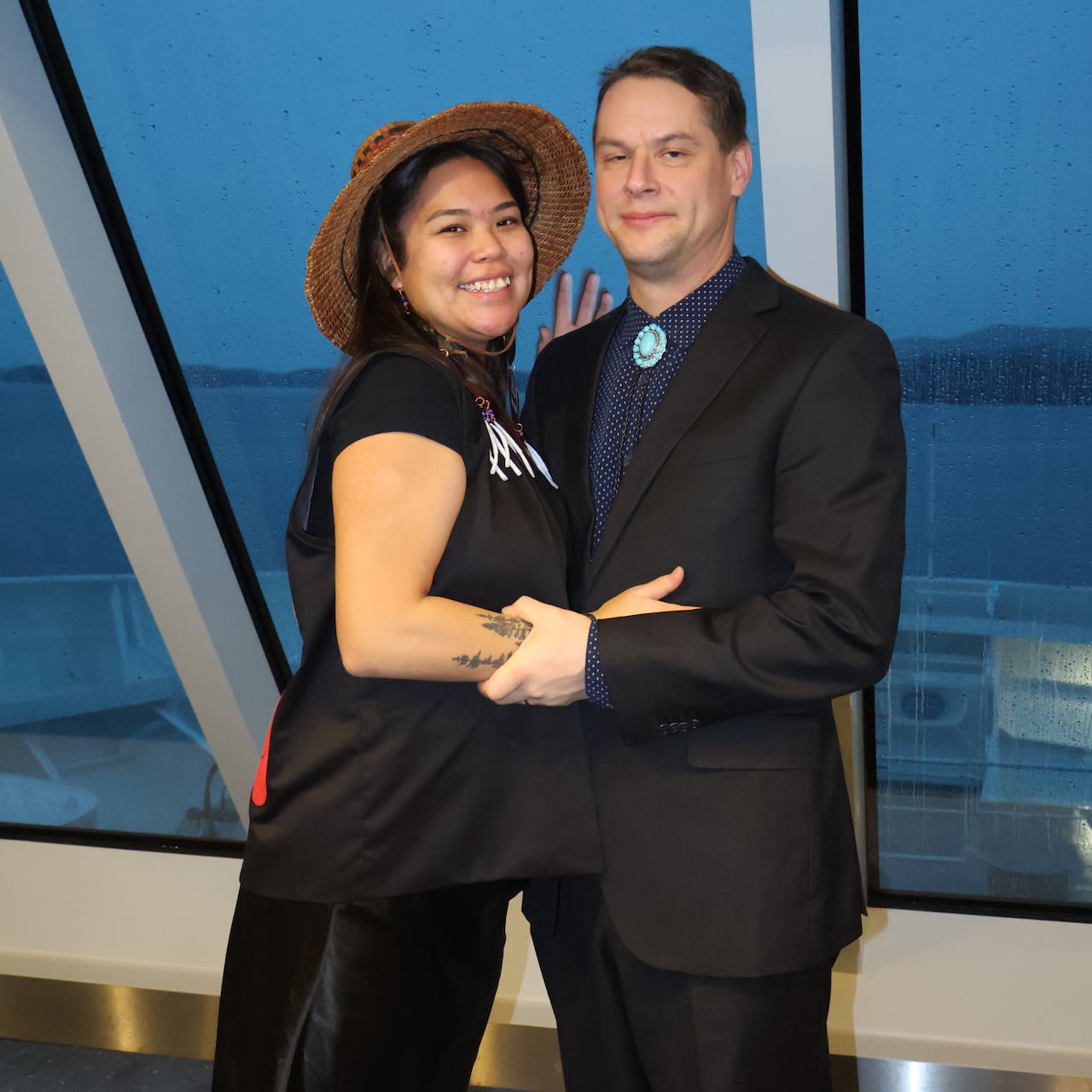 A pregnant women wearing a cedar hat and a man smile and hold each other with an oceanview in the background.