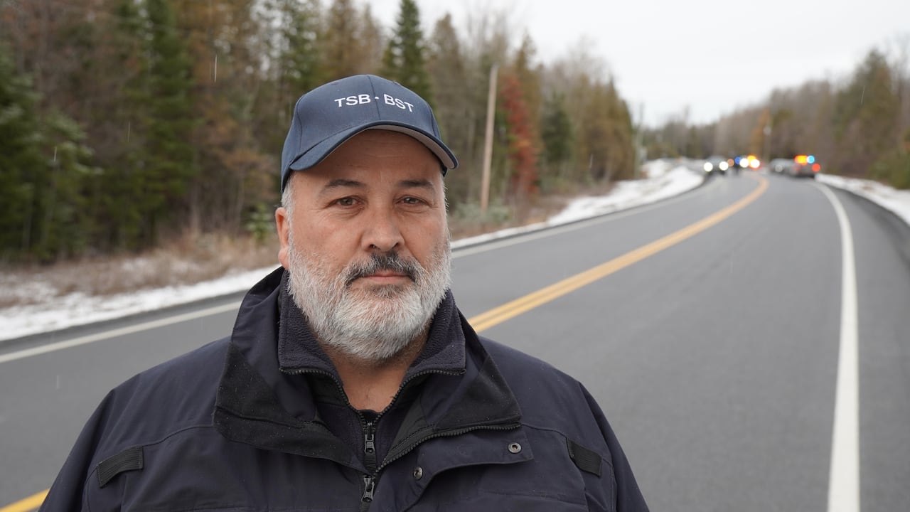 A man in a baseball cap stands on a highway in winter, with several police vehicles in the background.