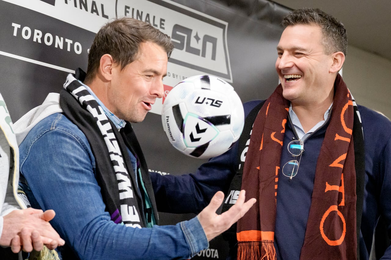 Two men draped in soccer scarves smile and throw a soccer ball.