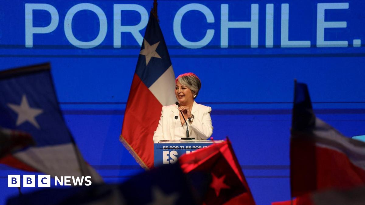Jeanette Jara stands at a podium with a blue backdrop, flanked by a Chilean flag.