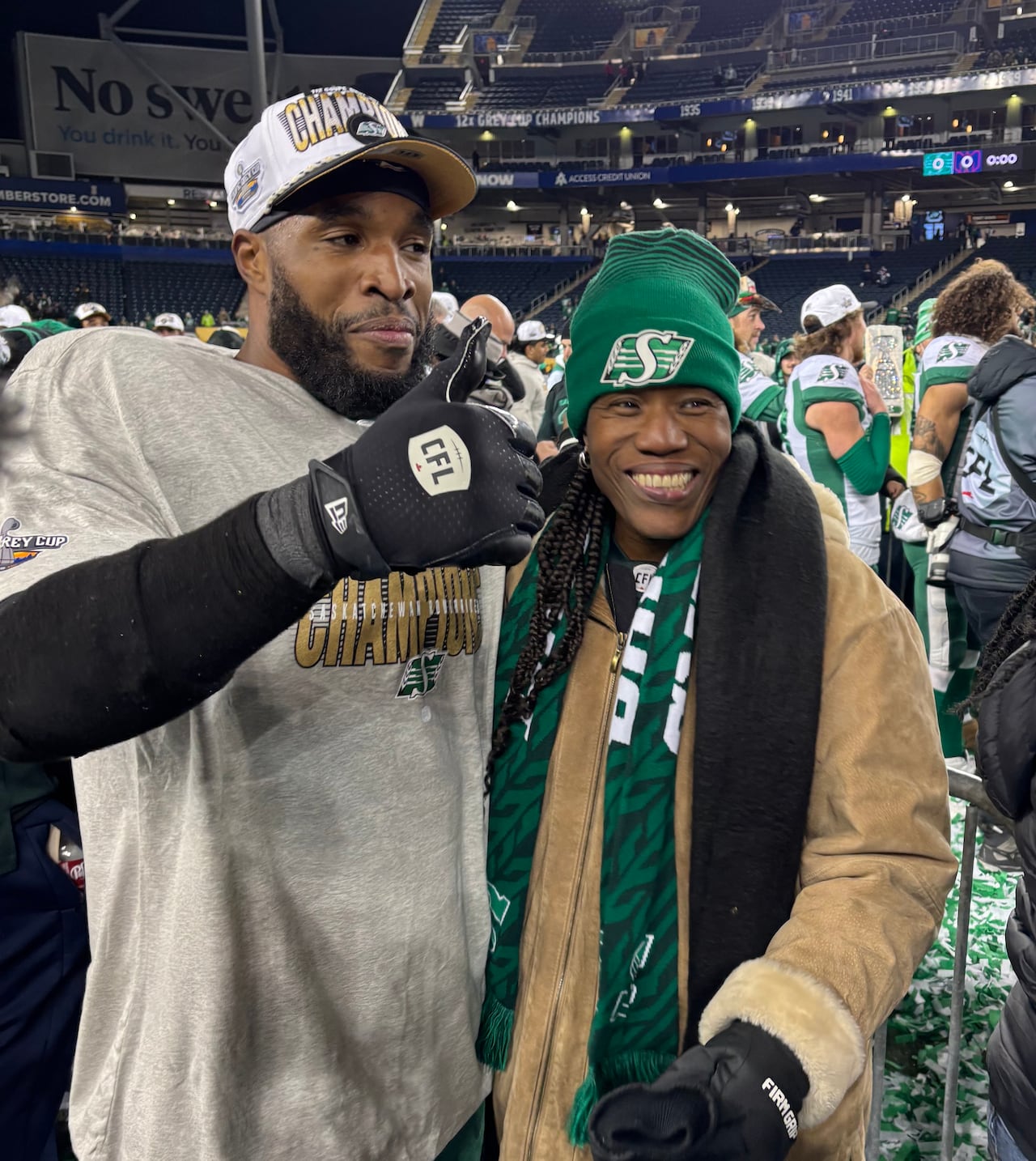 A man and a woman stand on a football field smiling. The man is wearing a championship hat and the woman wears a toque supporting the football team.