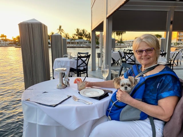 A woman and her dog sit at a table beside the water.