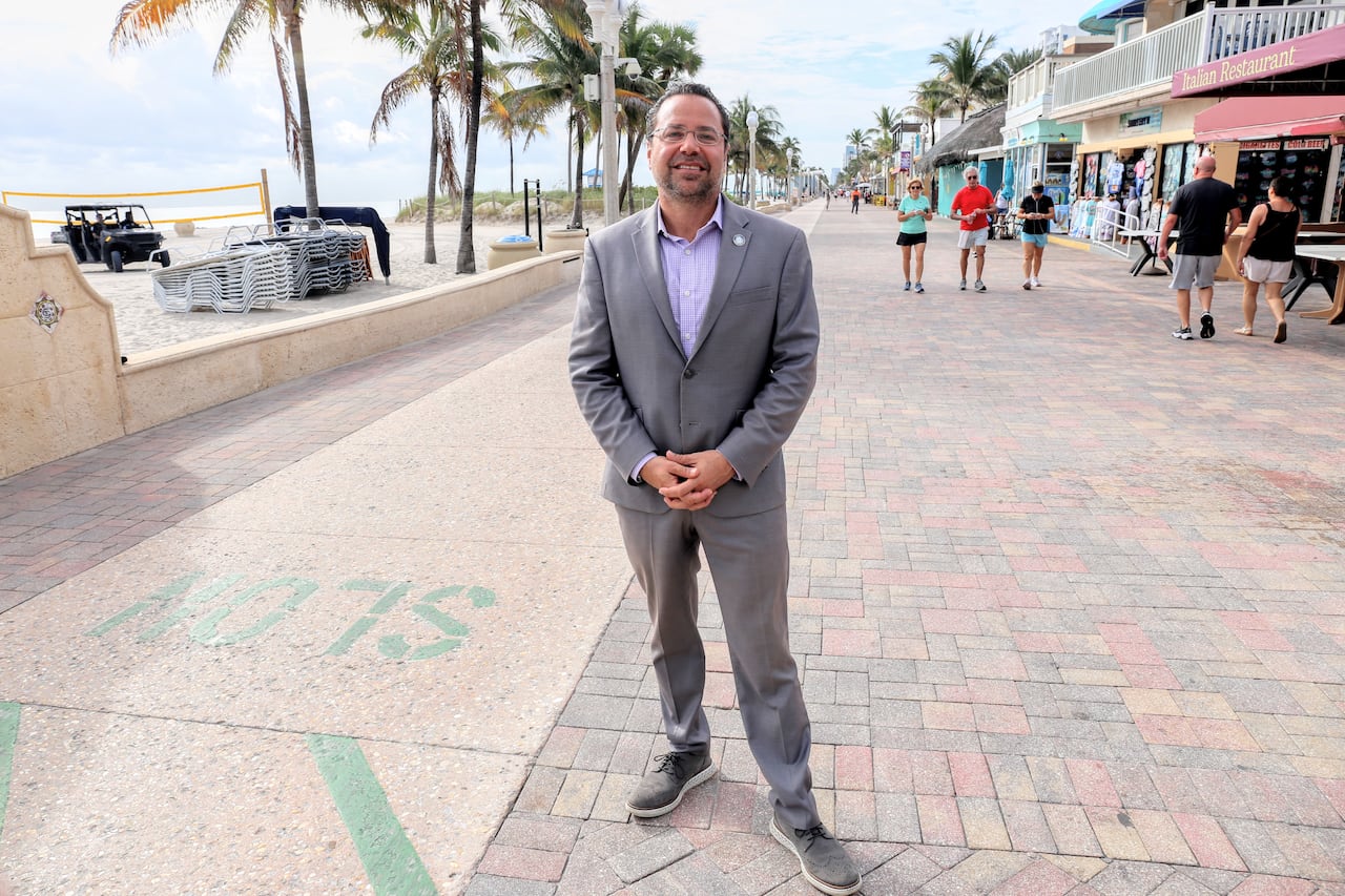 A man in a grey suit stands on a boardwalk by the beach.