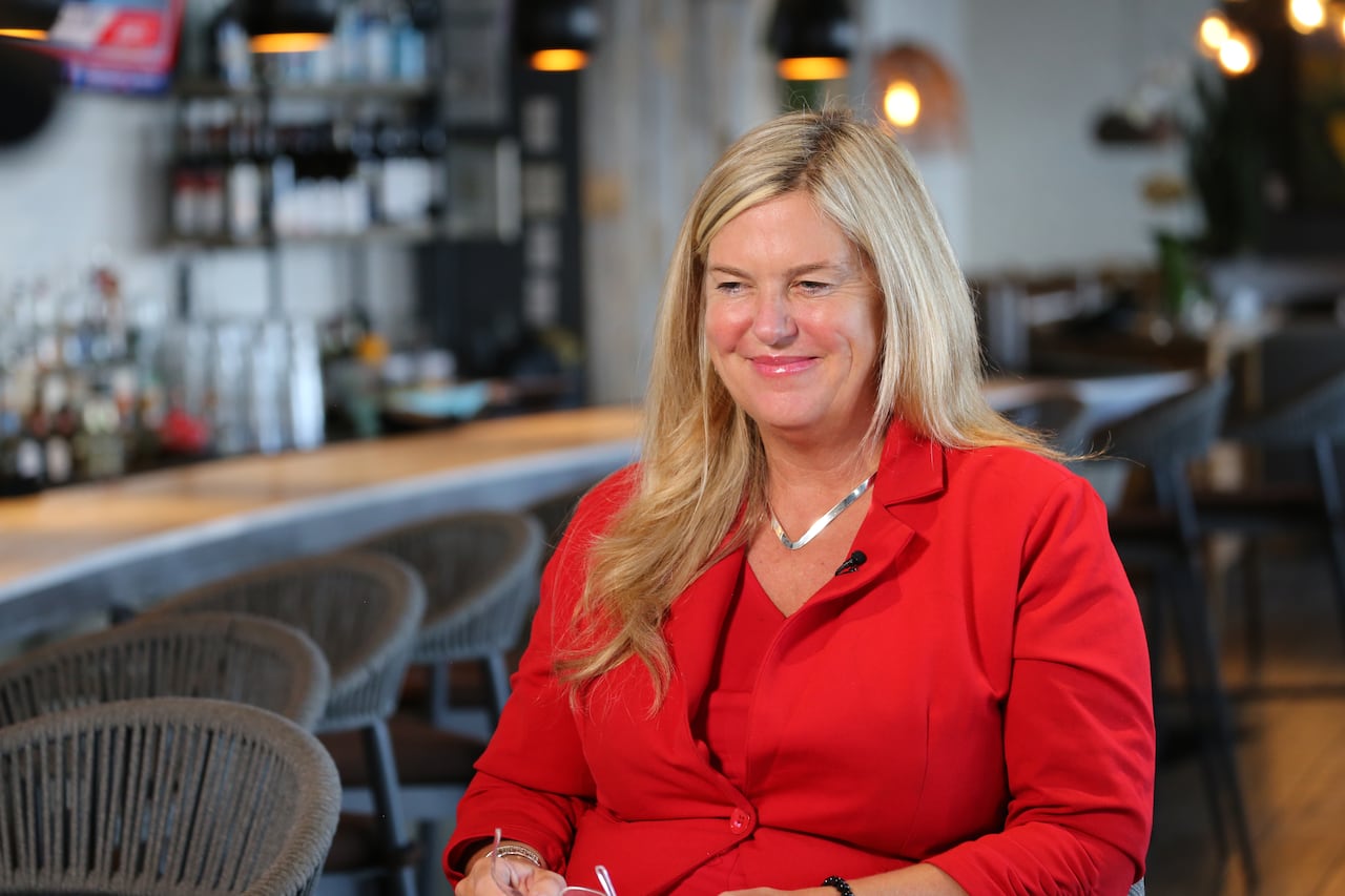 A woman in a red suit sits beside a table.