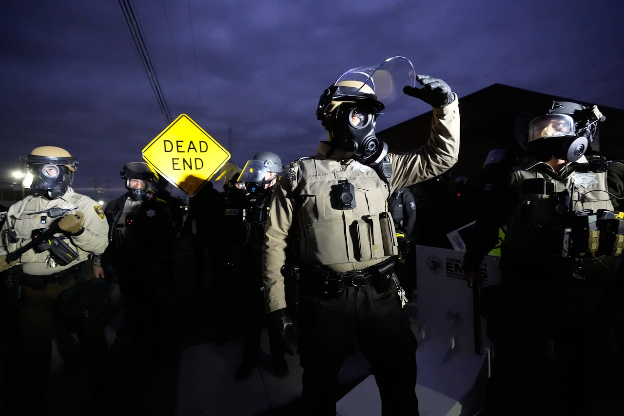 A handful of people with gas masks and bulletproof vests walk outside in a nighttime scene.