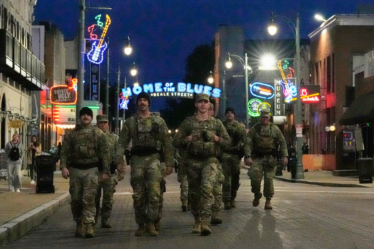 Several people in camouflage uniforms and carrying weapons walk down a city street that has buildings with neon signs.