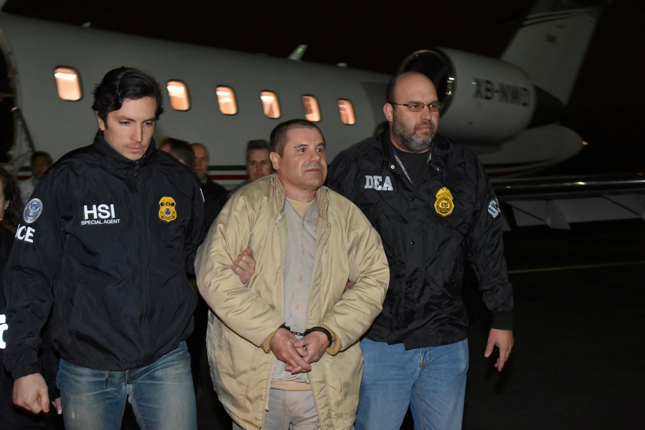 Man in light coloured clothes being escorted by two law official agents from an airplane