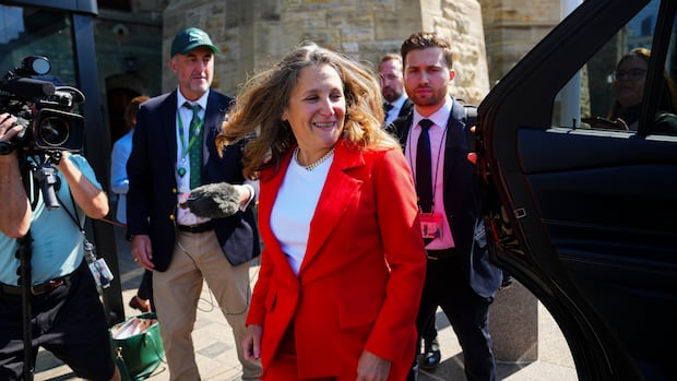 A man in a suit and a woman in a suit walk down a hallway with cameras following them.