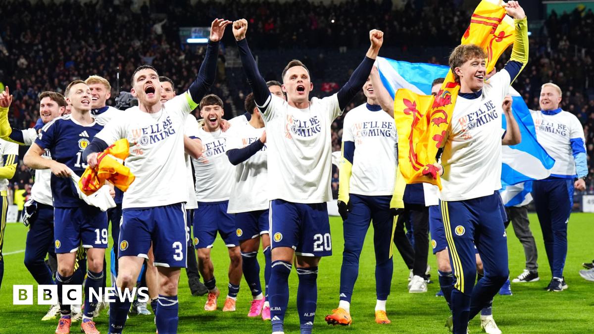 Football players celebrating on the pitch, wearing white shirts over their team kits and holding Scottish flags.