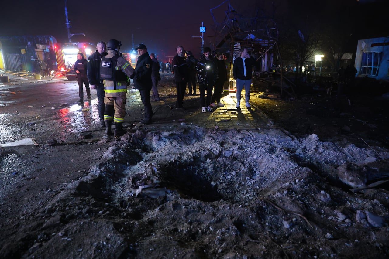 A nighttime scene shows first responders in an urban setting near a huge crater and other debris.