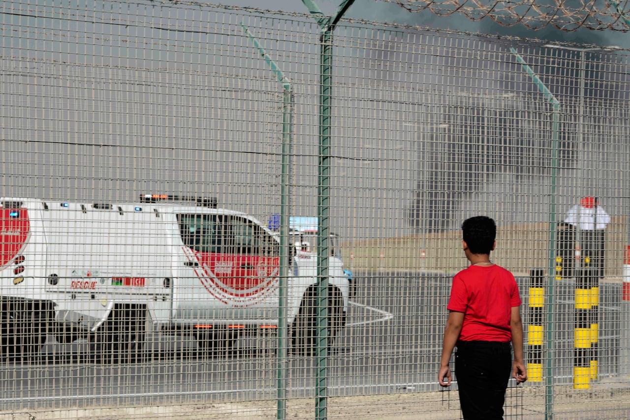 Person watching smoke billowing in the distance on a tarmac from behind a metal fence.