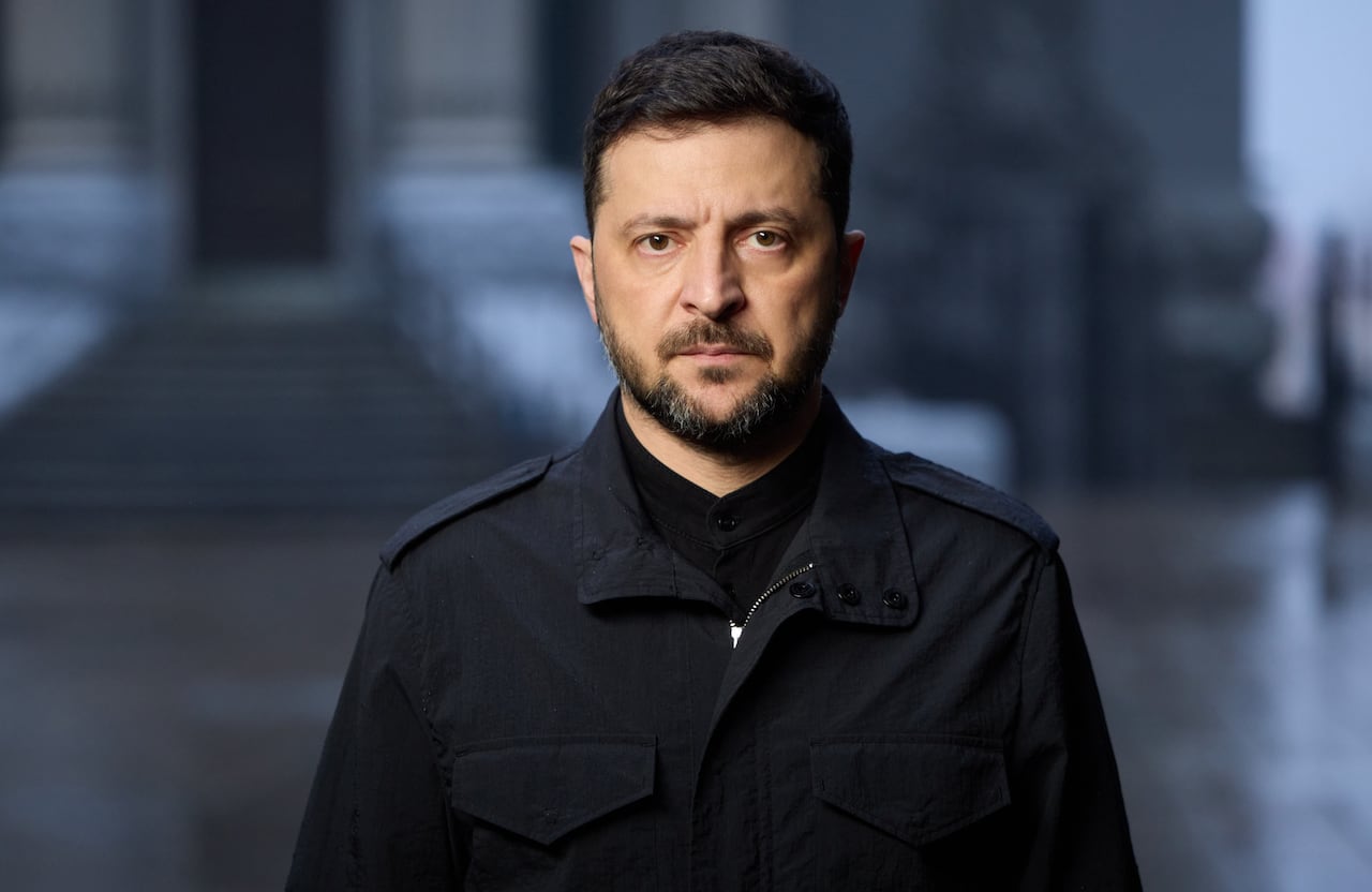 A man with black hair and a short beard, wearing a black jacket, stares into the camera as he stands in front of a building.